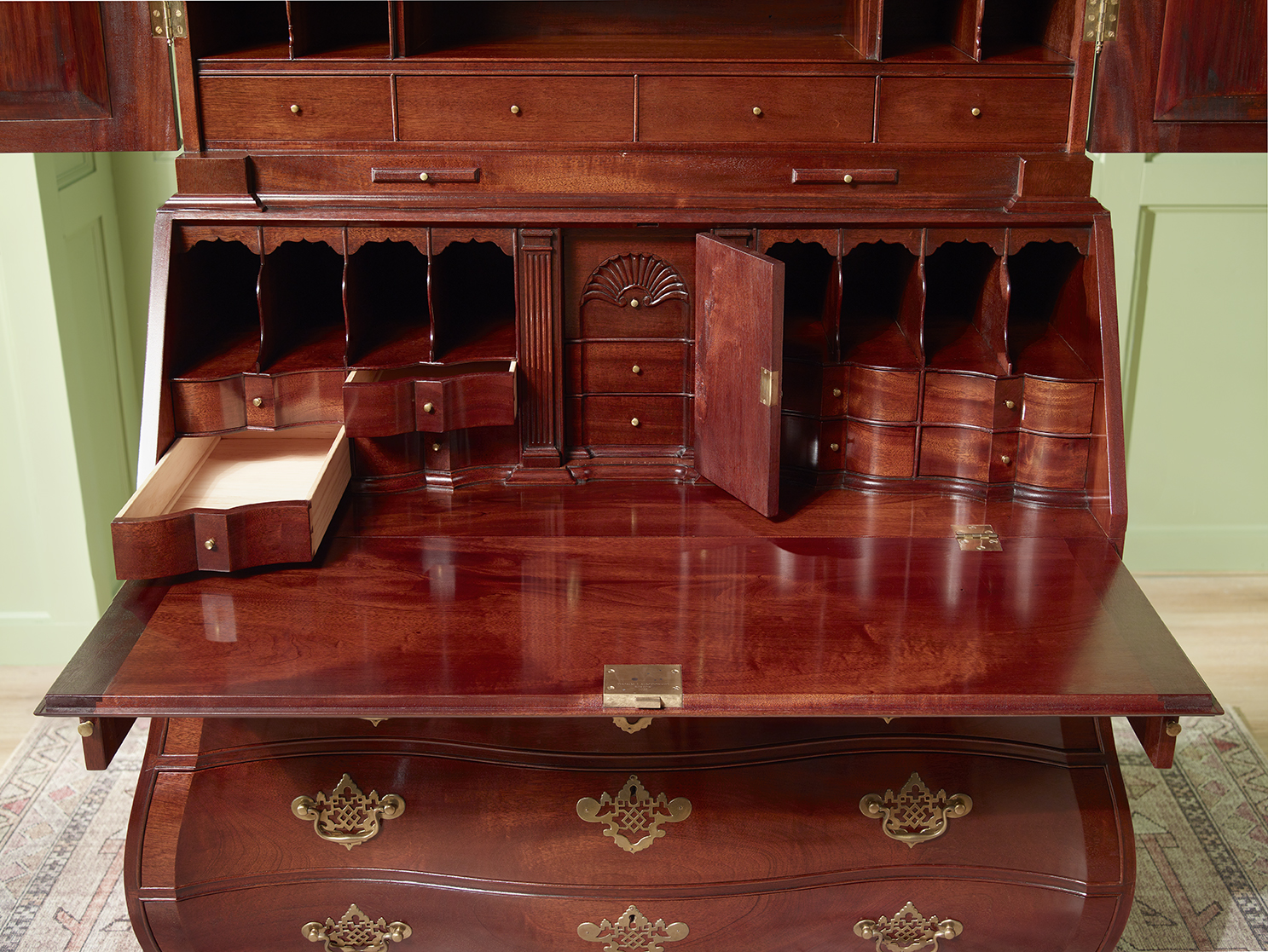 Open writing surface of a bombe secretary with scalloped drawers, pigeonholes, hidden compartments, and brass hardware.