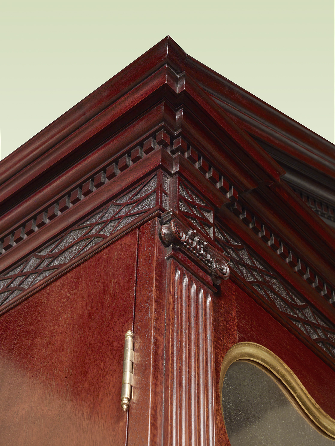 Corner view of a mahogany cabinet showing carved dentil molding, fluted pilaster, lattice pattern carving, and gilded glass panel edge.