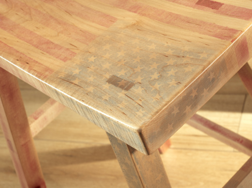 Image show the top corner view of a rectangular maple stool with through wedged mortis and tenons and four splayed legs. The top is adorned with a distressed American flag design and there are faded Stars and Stripes going down the legs.