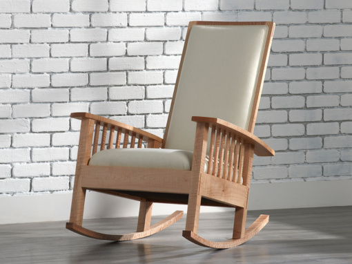 An arts & crafts style figured maple rocking chair upholstered with a soft cream colored wool fabric shown in front of a white subway tile wall with gray grout on a polished cement floor.