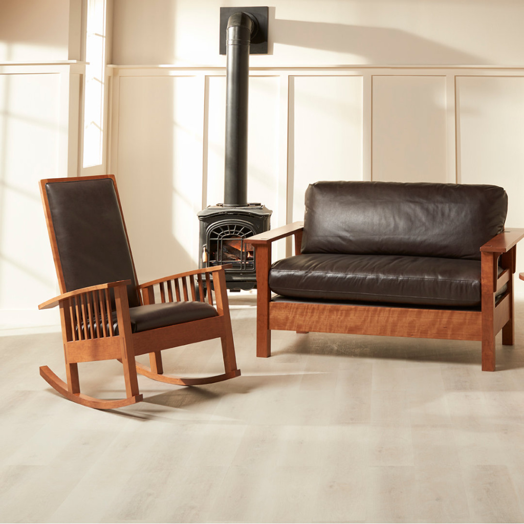 Square A plush, handmade leather upholstered cherry Loveseat next to a cherry rocking chair with the same dark, peppercorn colored leather upholstery sit in a bright room with a wood-burning stove in the background.