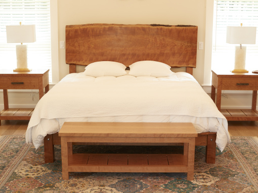 a live edge cherry bed shown in a bright bedroom with white linens. a cherry nightstand at either side of the bed and a long cherry wood bench.