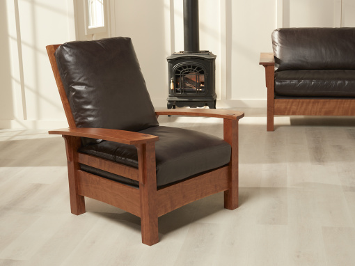 A Morris style adjustable back arm chair made of natural cherry and peppercorn colored Italian leather cushions in a bright room with a wood burning stove and matching loveseat in the background.