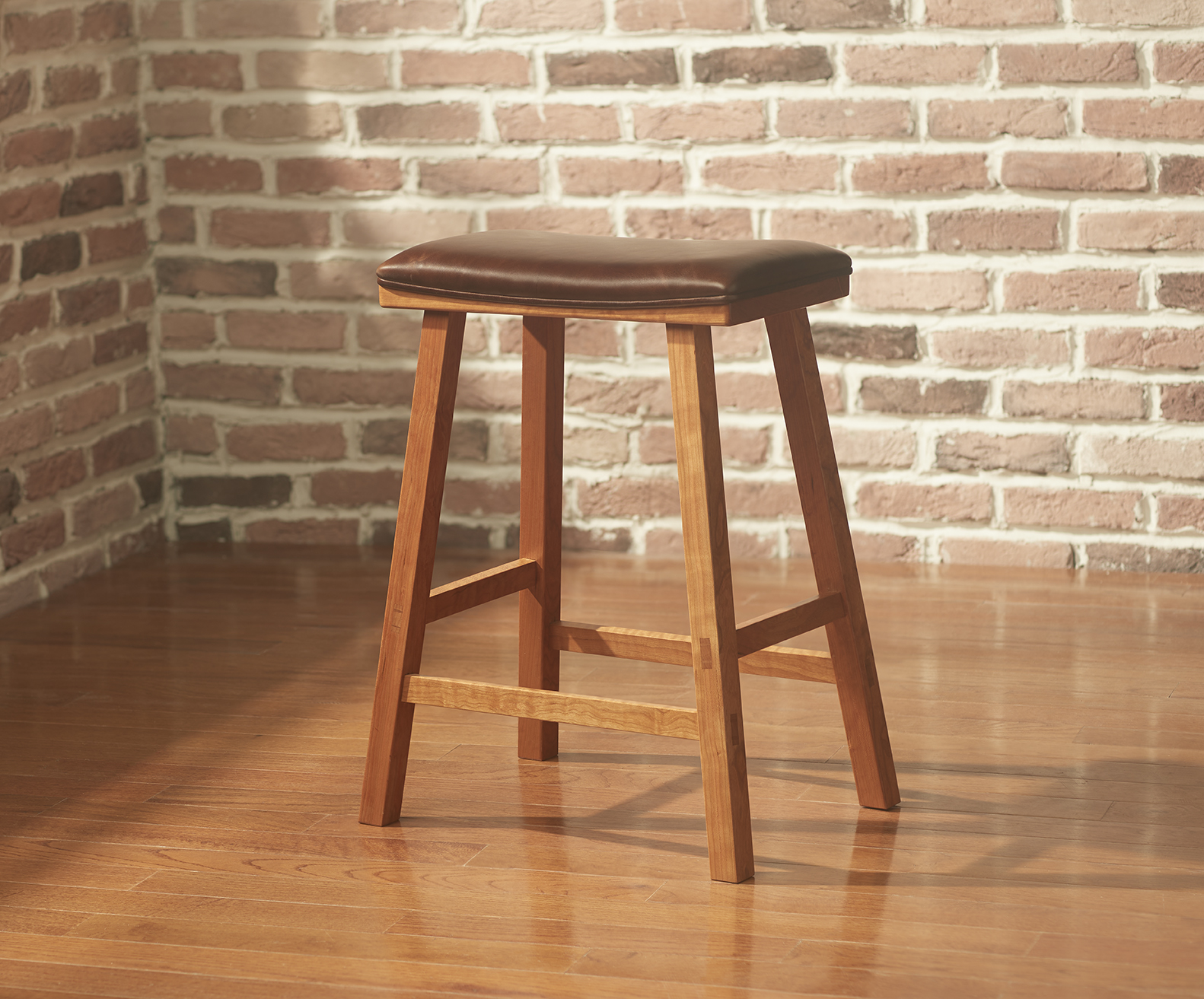 A simple cherry bar stool with a rectangular seat and squared splayed legs with chocolate brown leather upholstery in a room with exposed brick walls and hardwood floor.