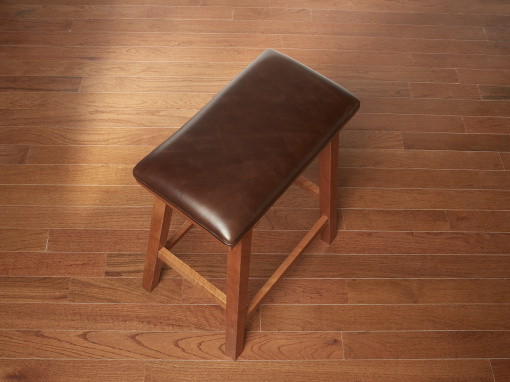 A top angled view of a rectangular cherry barstool with a brown leather seat and squared splayed legs shown on a hardwood floor.
