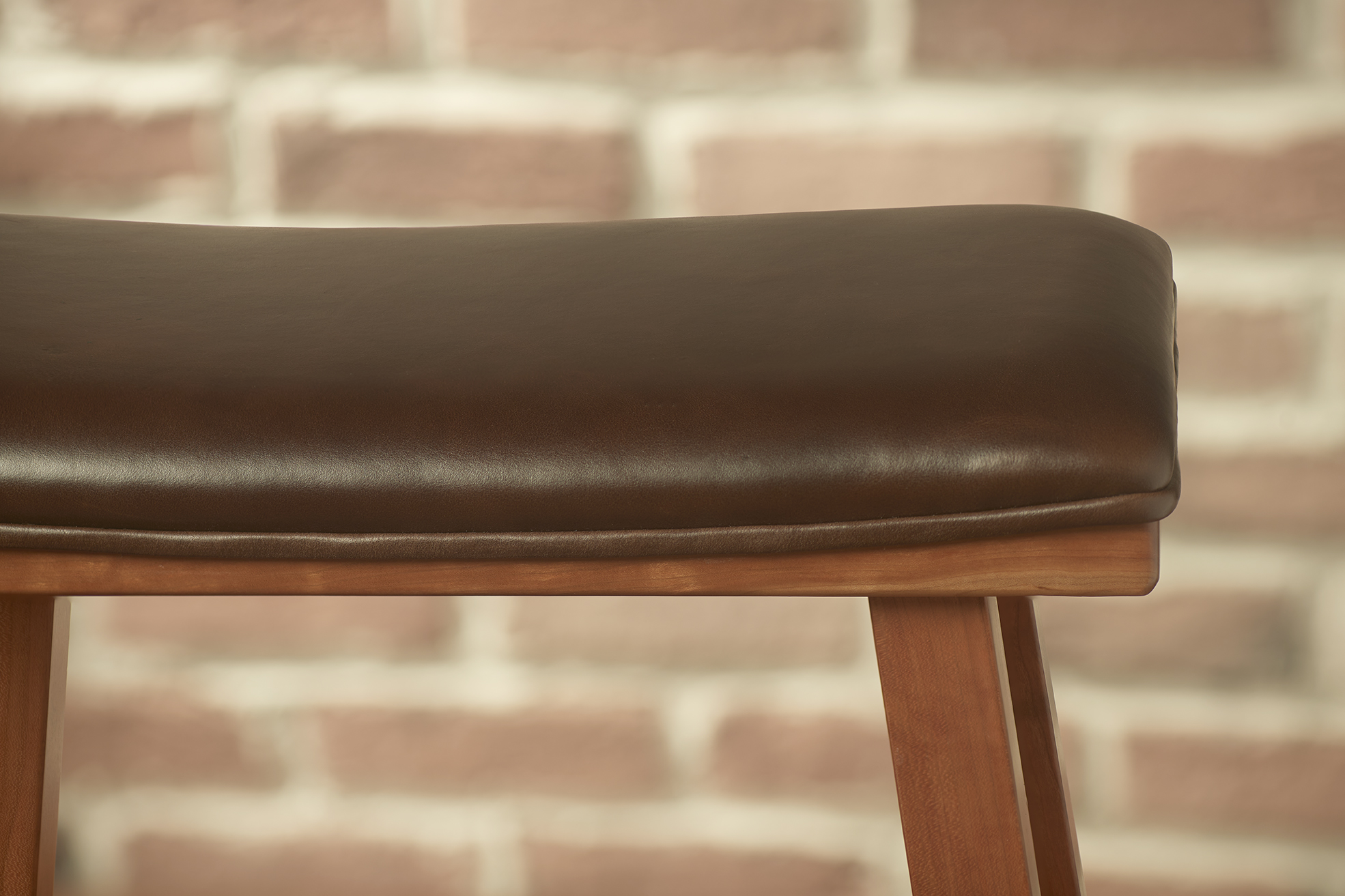 A close up side view of an upholstered cherry kitchen barstool showing the brown leather seat with its piping around the curve of the seat.