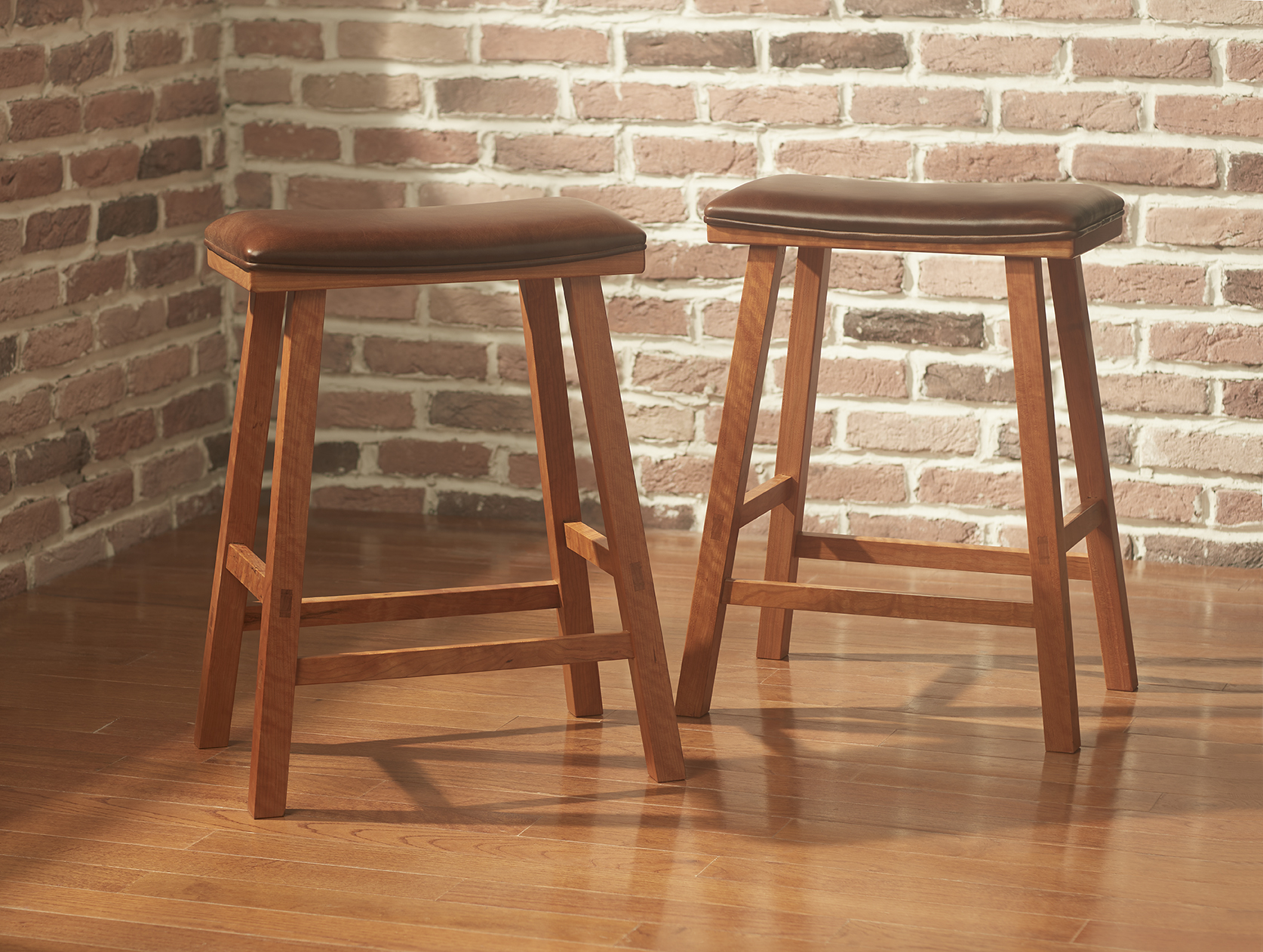 A pair of rectangular upholstered Cherry Kitchen Barstools with brown leather seats and squared splayed legs shown side by side in a room with an exposed brick wall and a hardwood floor.