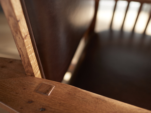 Close-up of exposed mortise and tenon joinery on the back of a handcrafted oak rocking chair arm, with brown leather seat and backrest partially visible.